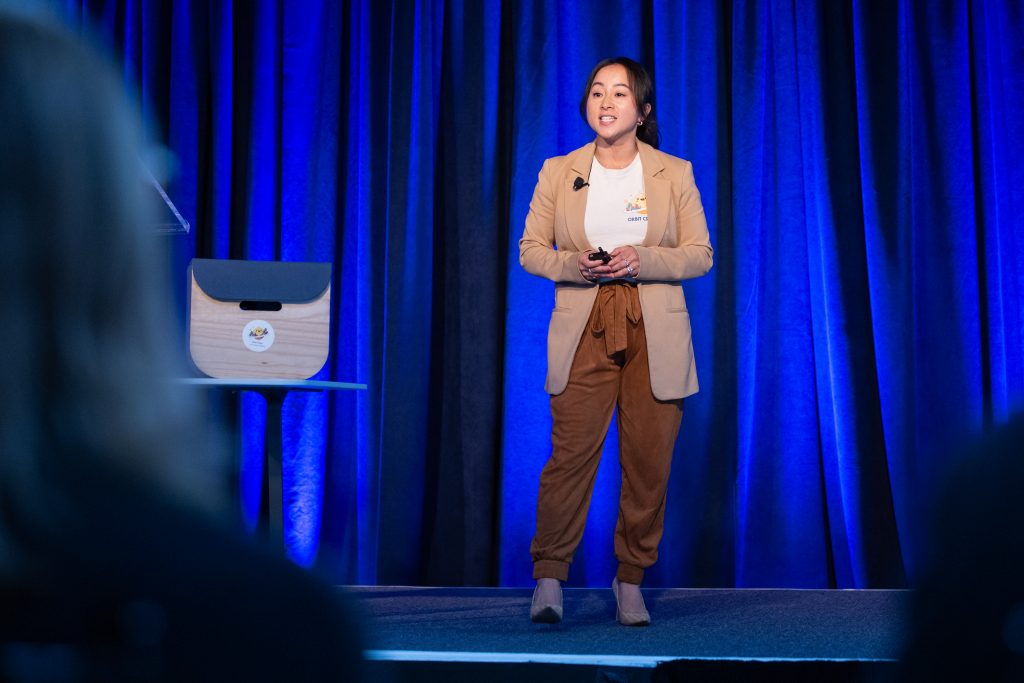 A woman in brown business attire stands on a stage in front of blue curtains.