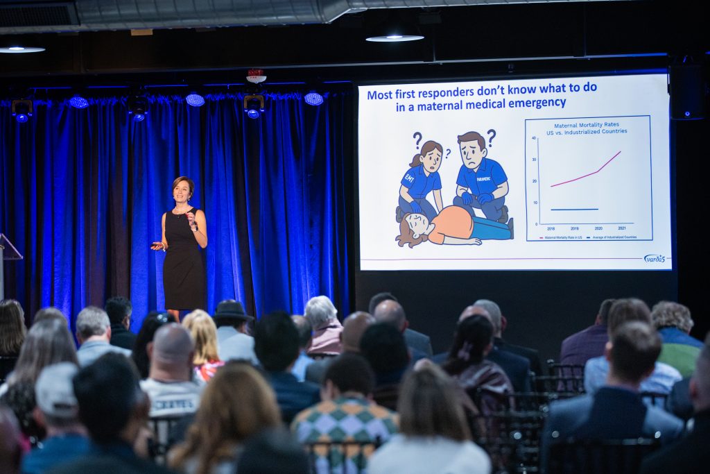 A woman addresses a crowd from a stage in front of a screen that says "Most first responders don't know what to do in a maternal medical emergency."