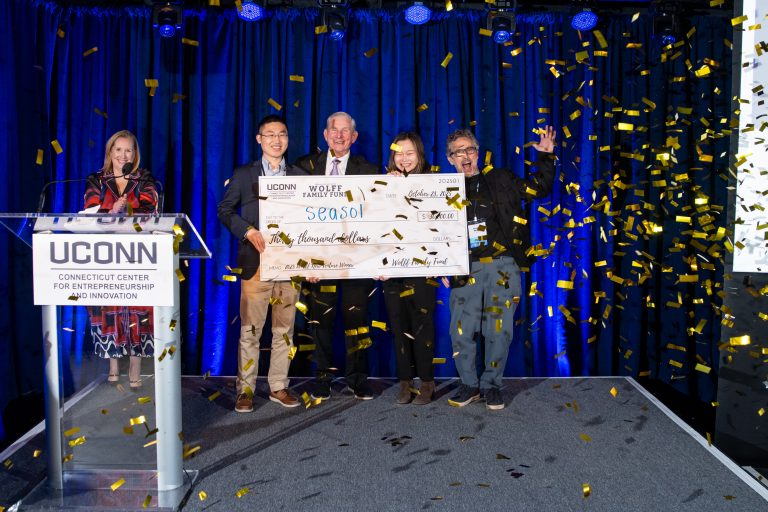 A woman stands behind a white podium while a group behind her holds a large novelty check as confetti falls.