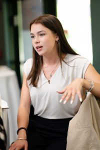 A young woman in a white blouse sits in a chair and speaks.