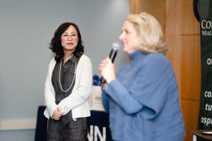 A woman in a white sweater watches as a woman in blue speaks into a microphone to a crowd not in the frame.