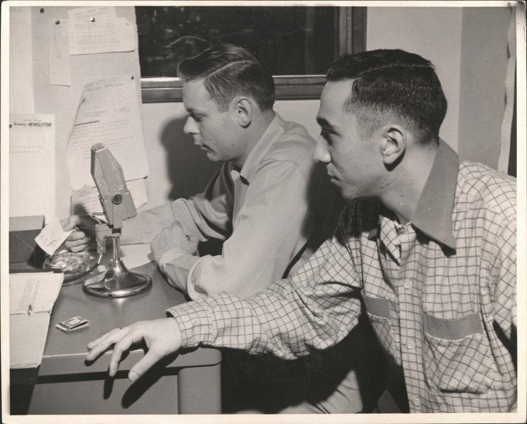 A black and white archival photo shows two students at a radio desk.