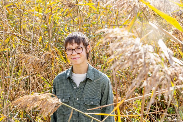 A student in a grey shirt stands in a field of phragmites, a tall, pale brown grass.