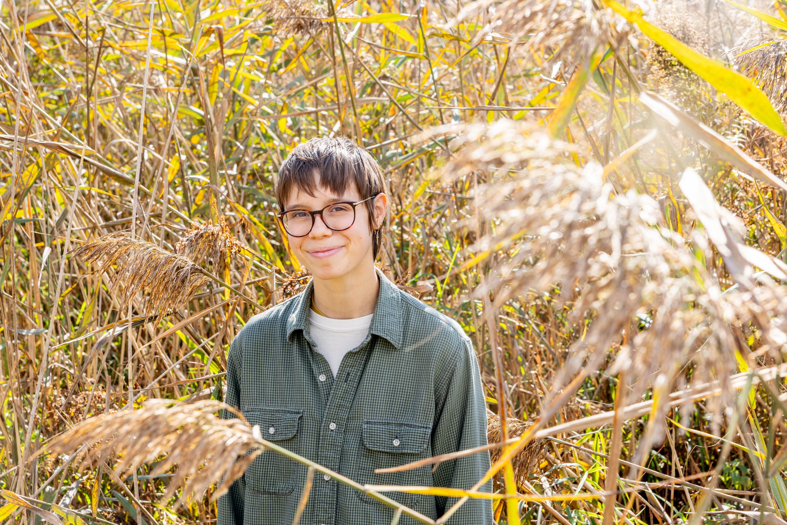 A student in a grey shirt stands in a field of phragmites, a tall, pale brown grass.