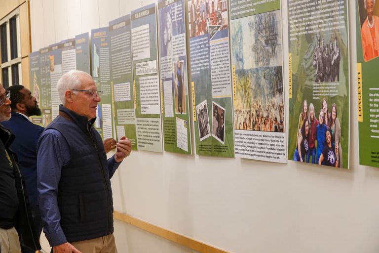 Three visitors examine poster displays at the Bloomfield Mosaic exhibition wall.