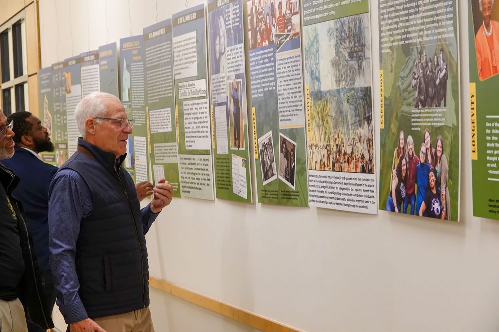 Three visitors examine poster displays at the Bloomfield Mosaic exhibition wall.