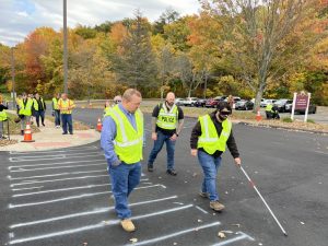 People wearing reflective vests participate in an outdoor ADA accessibility training, with one person using a white cane to simulate visual impairment.