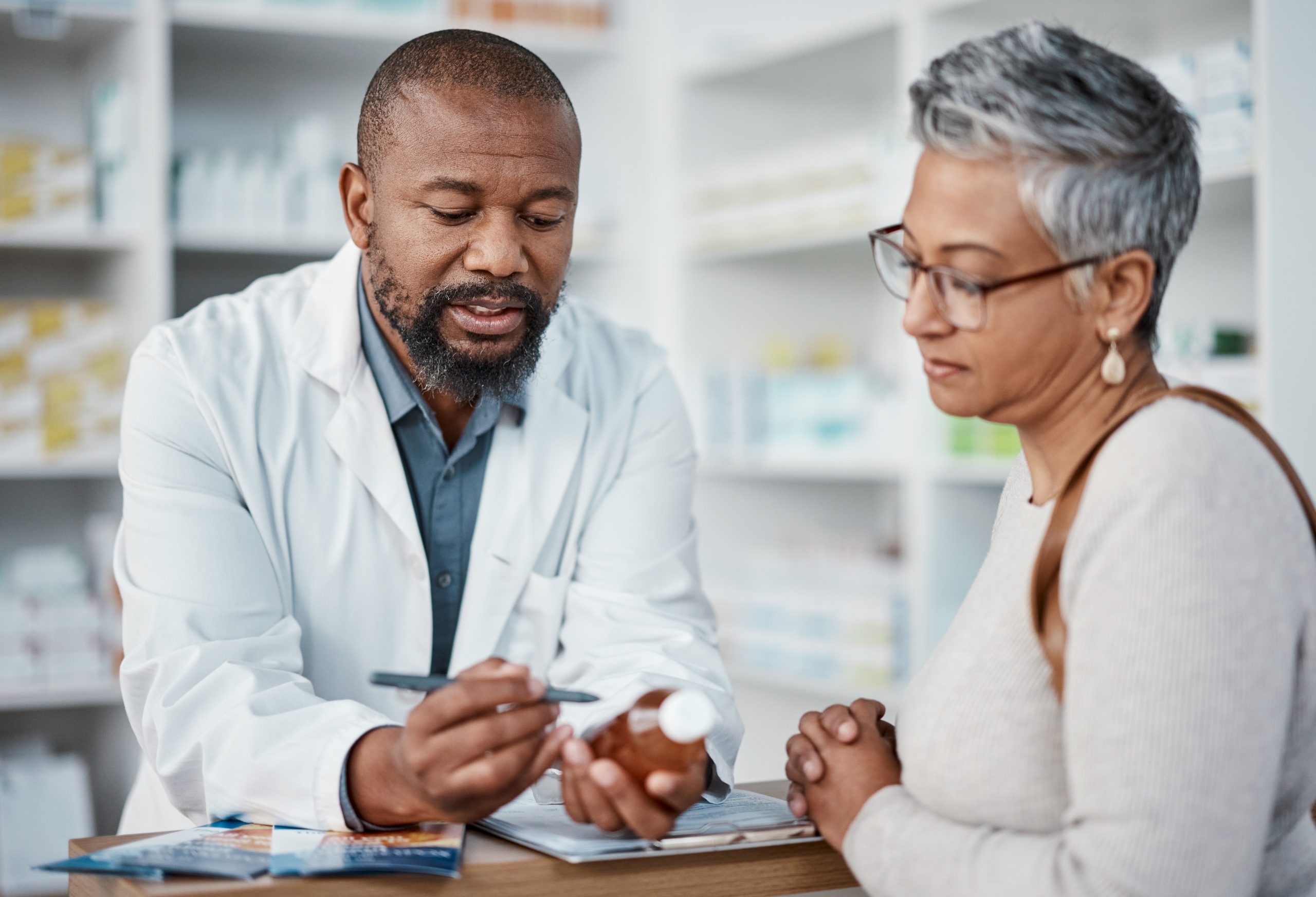 A pharmacist in a white coat examines the label on a prescription bottle with a patient.