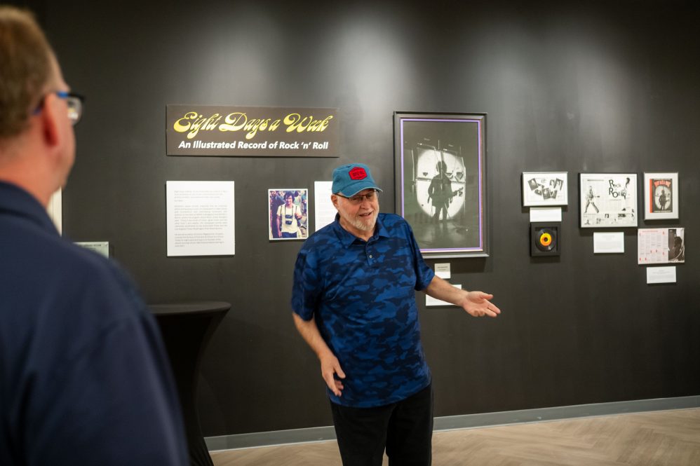 A man stands in front of a wall with photos and rock and roll memorabilia, gesturing to a tour group.
