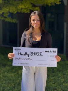 A young woman holds a large presentation check that says "HuskySHARE" on it.