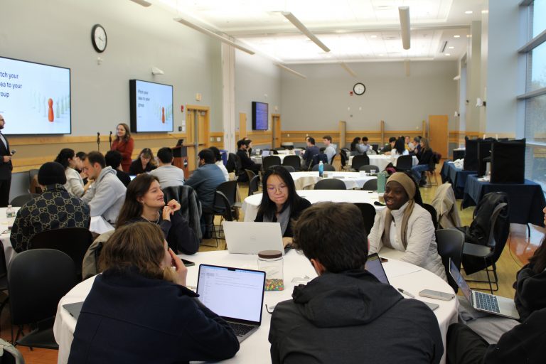Groups of students work together at round tables during an interactive workshop session in a large event room.
