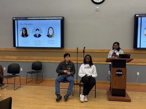 Two student panelists sit on stage while a moderator speaks at a podium during a UConn event, with a screen displaying panelist names and photos behind them.