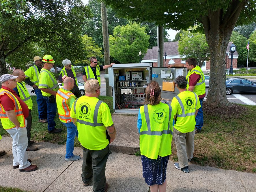 A group of public works employees in safety vests gather outdoors around an open traffic signal cabinet for a training demonstration.