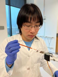 A person in a lab coat and purple gloves places a dyed water droplet into a ribbon array.
