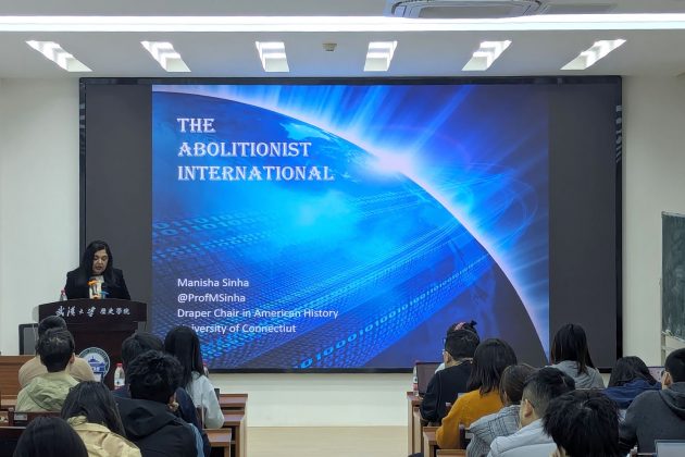 Woman stands at the front of a room behind podium, in front of an audience. 