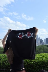 A person holds up a black t-shirt with graphics and the word "Ochi" printed on it.