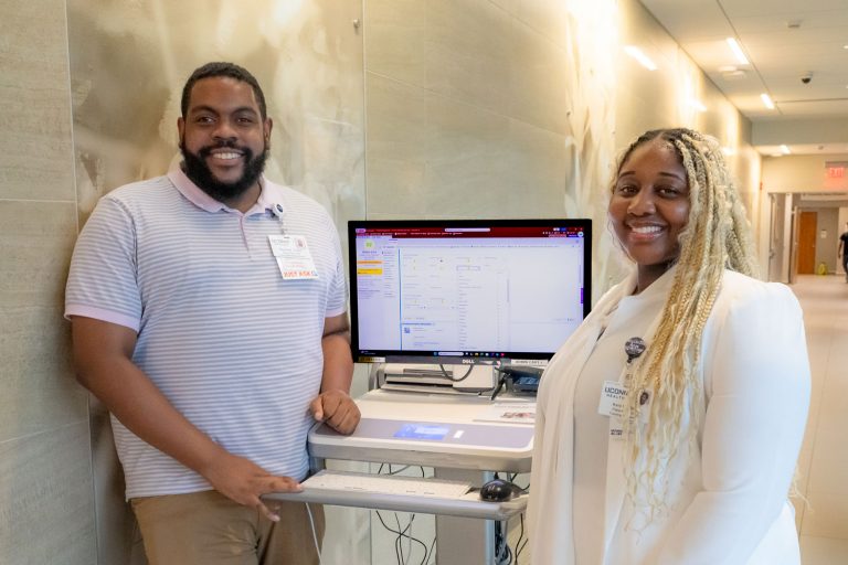 portrait man and woman stand beside a computer workstation used for patient registration training.
