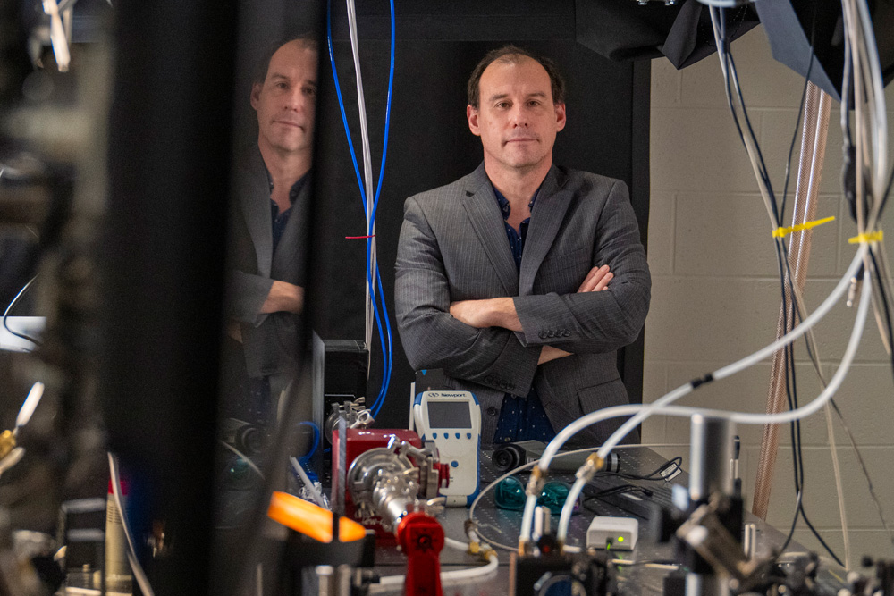 A white man in a grey jacket stands with his arms crossed in a laboratory.