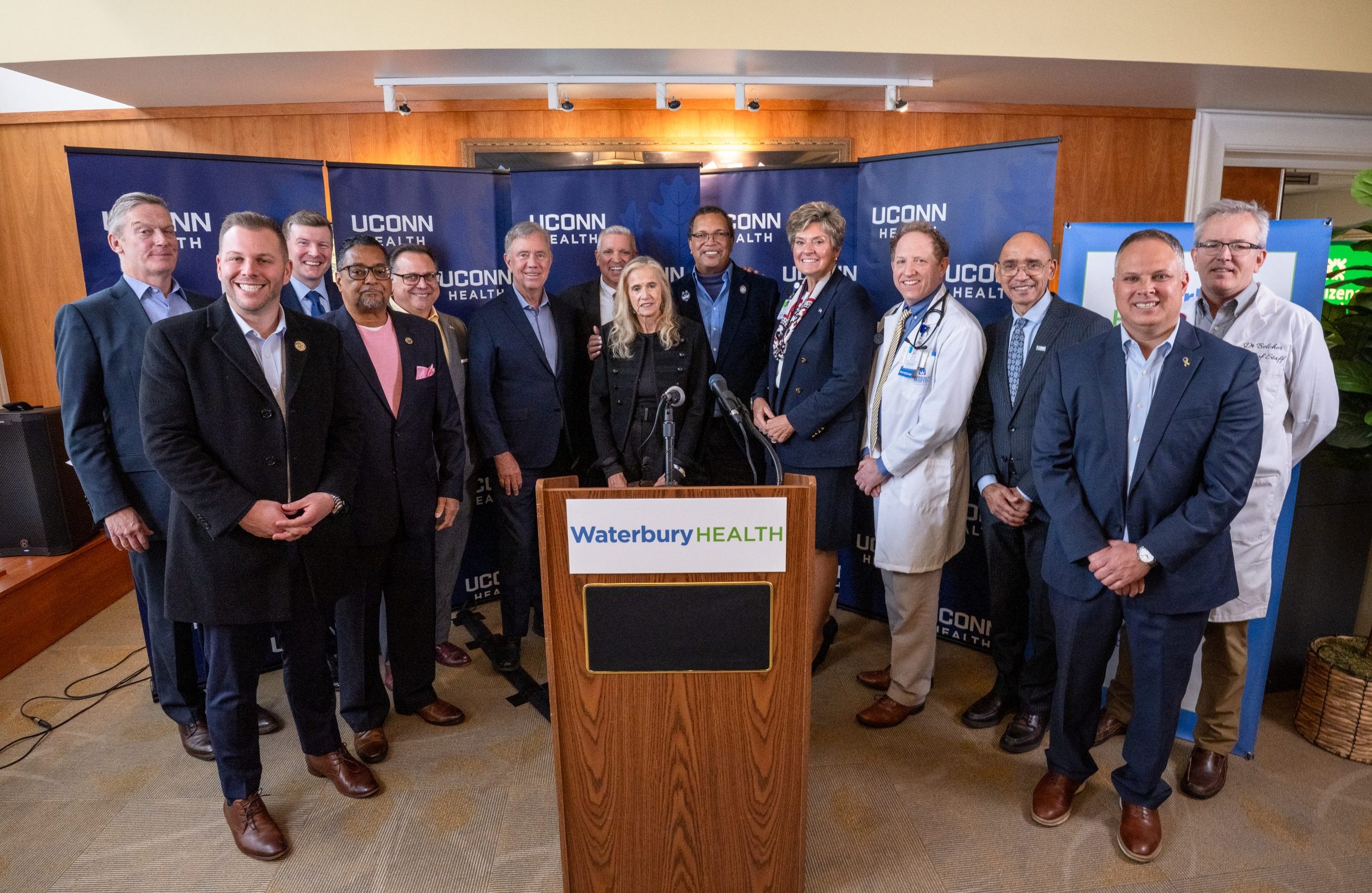 Several men and women including legislators, hospital leaders and the governor standing behind a Waterbury HEALTH podium and infront of UConn Health Backdrop