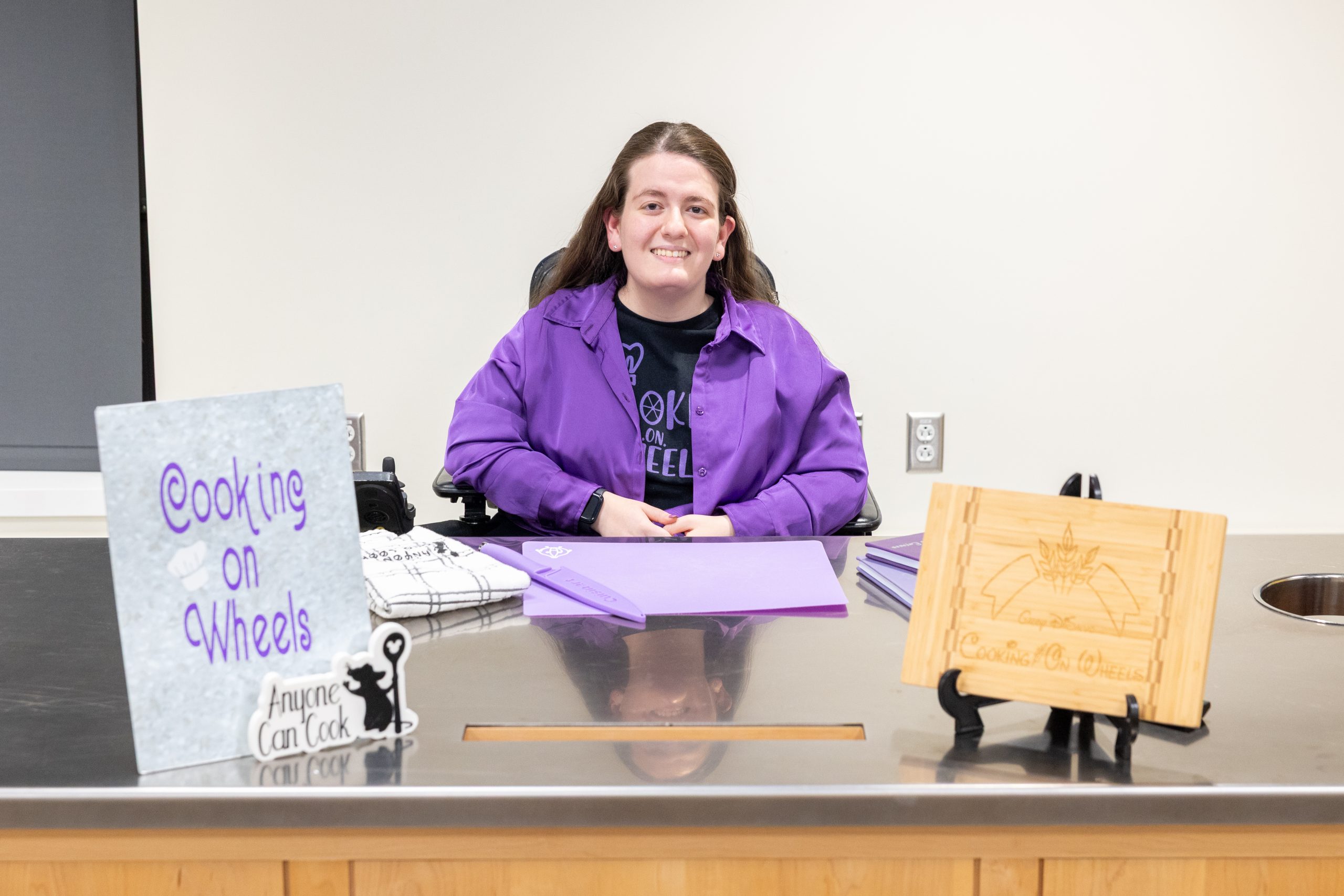 A girl in a purple jacket sits at a table with a book and a cutting board propped up
