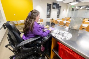 A girl in a purple jacket sits at a table cutting lettuce with a cloth that reads 'anyone can cook'