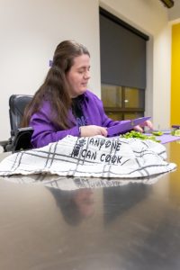 A girl in a purple jacket sits at a table cutting lettuce with a cloth that reads 'anyone can cook'