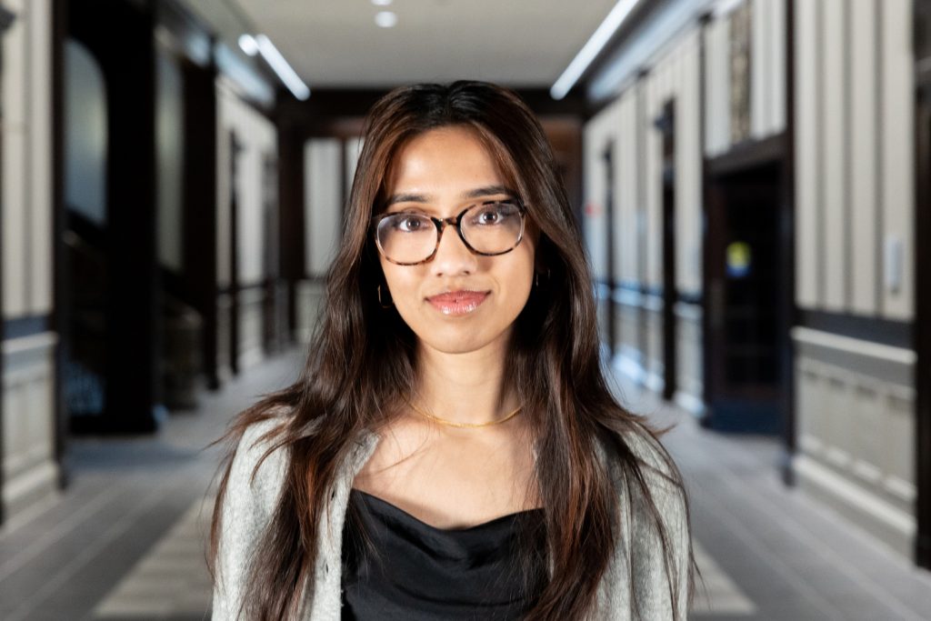 A woman with long brown hair and glasses poses for a photo in an academic building