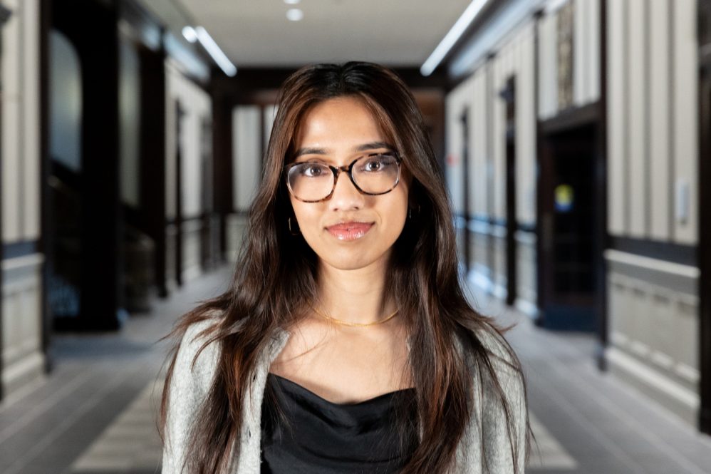 A woman with long brown hair and glasses poses for a photo in an academic building