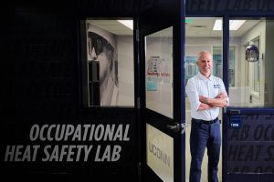 A man in a white shirt and blue pants stands in the doorway of a building. The sign outside reads "Occupational Heat Safety Lab"