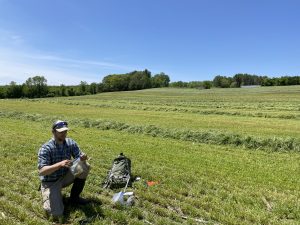A researcher kneeling down to collect samples in a green hayfield with trees in the background, and a cloudless blue sky above.