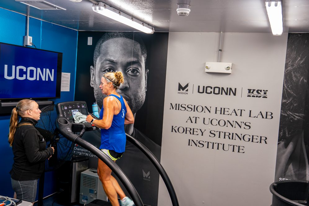 A woman runs on a treadmill while another woman stands next to it speaking to her. The wall behind them reads MISSION HEAD LAB AT UCONNS KOREY STRINGER INSTITUTE