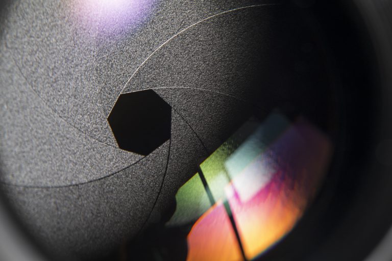 Macro view of a camera lens aperture with overlapping blades and colorful light reflections.