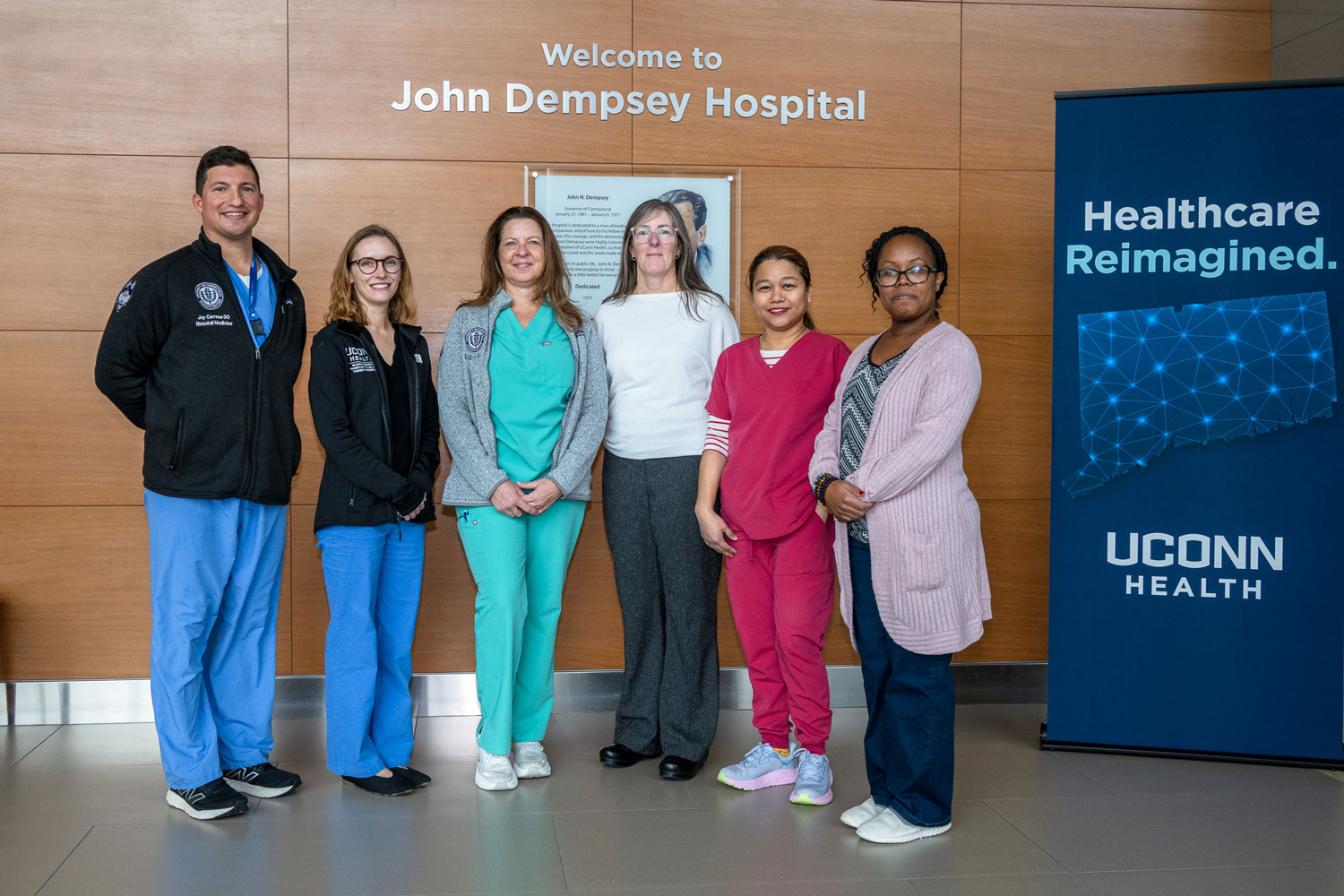 Group portrait, one man, five women, indoors in front of "Welcome to John Dempsey Hospital" sign and next to "Healthcare Reimagined" banner