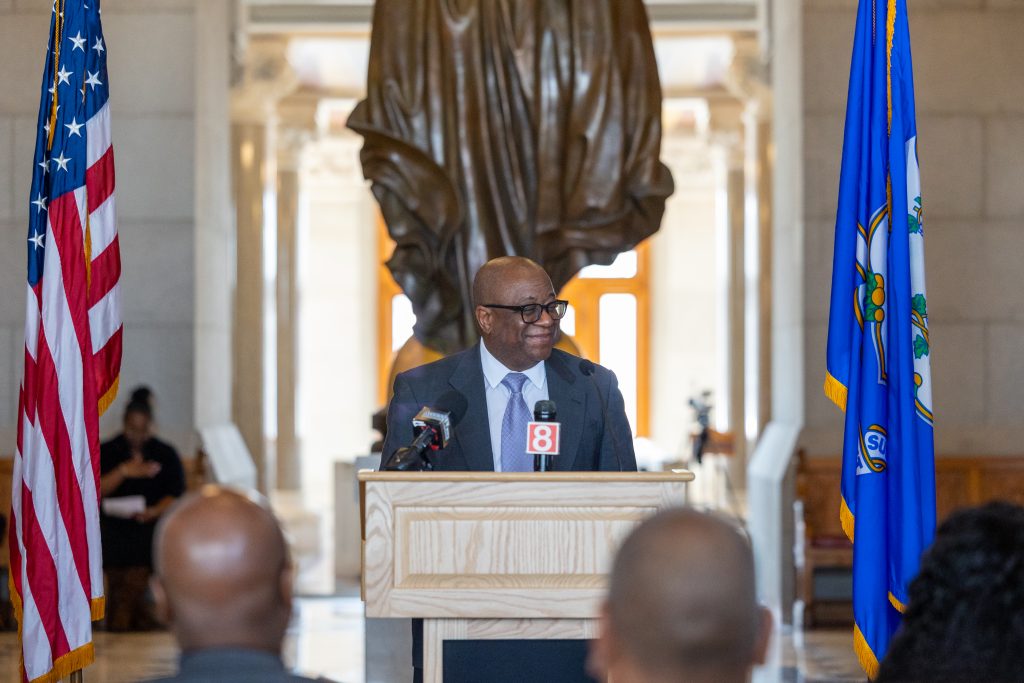A man stands at a podium flanked by US and Connecticut flags.