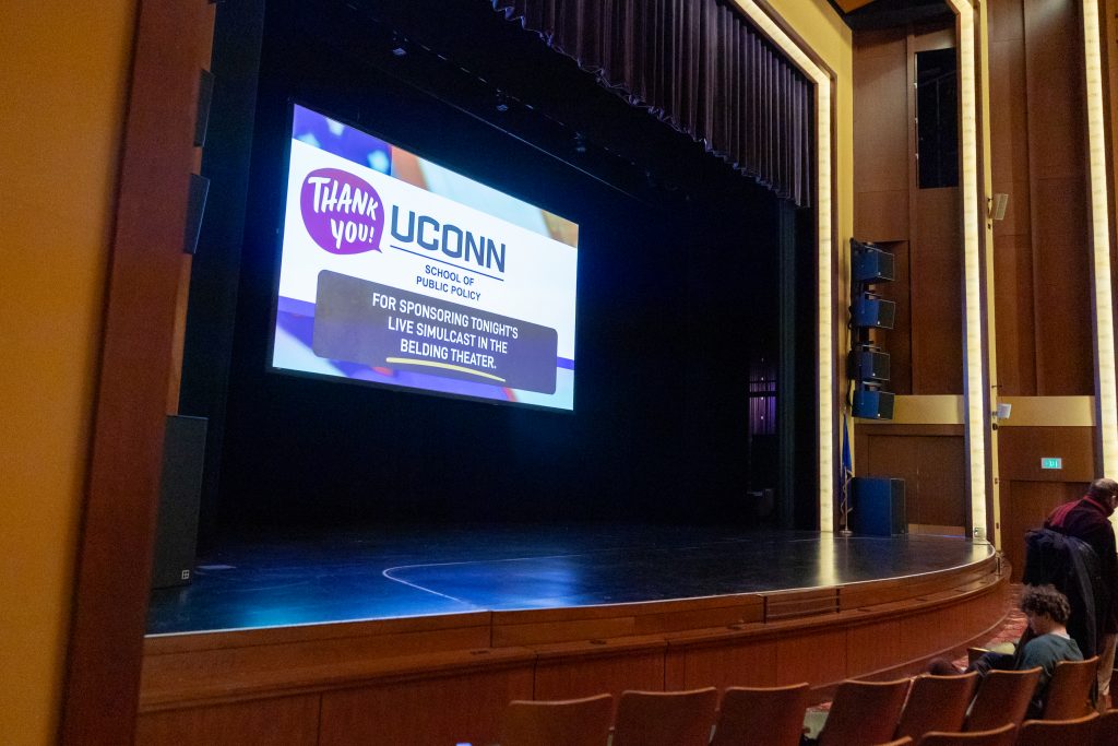 A screen above a stage thanks the UConn School of Public Policy for sponsoring an event.