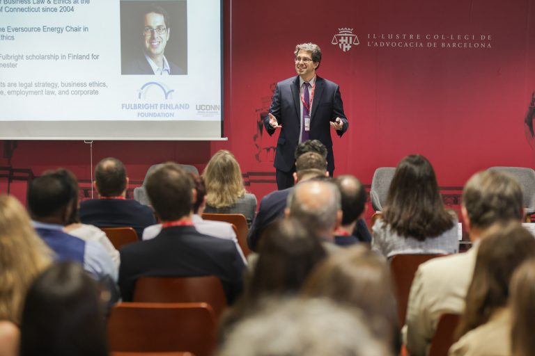 A man in a suit standing before a red background addresses a large audience.