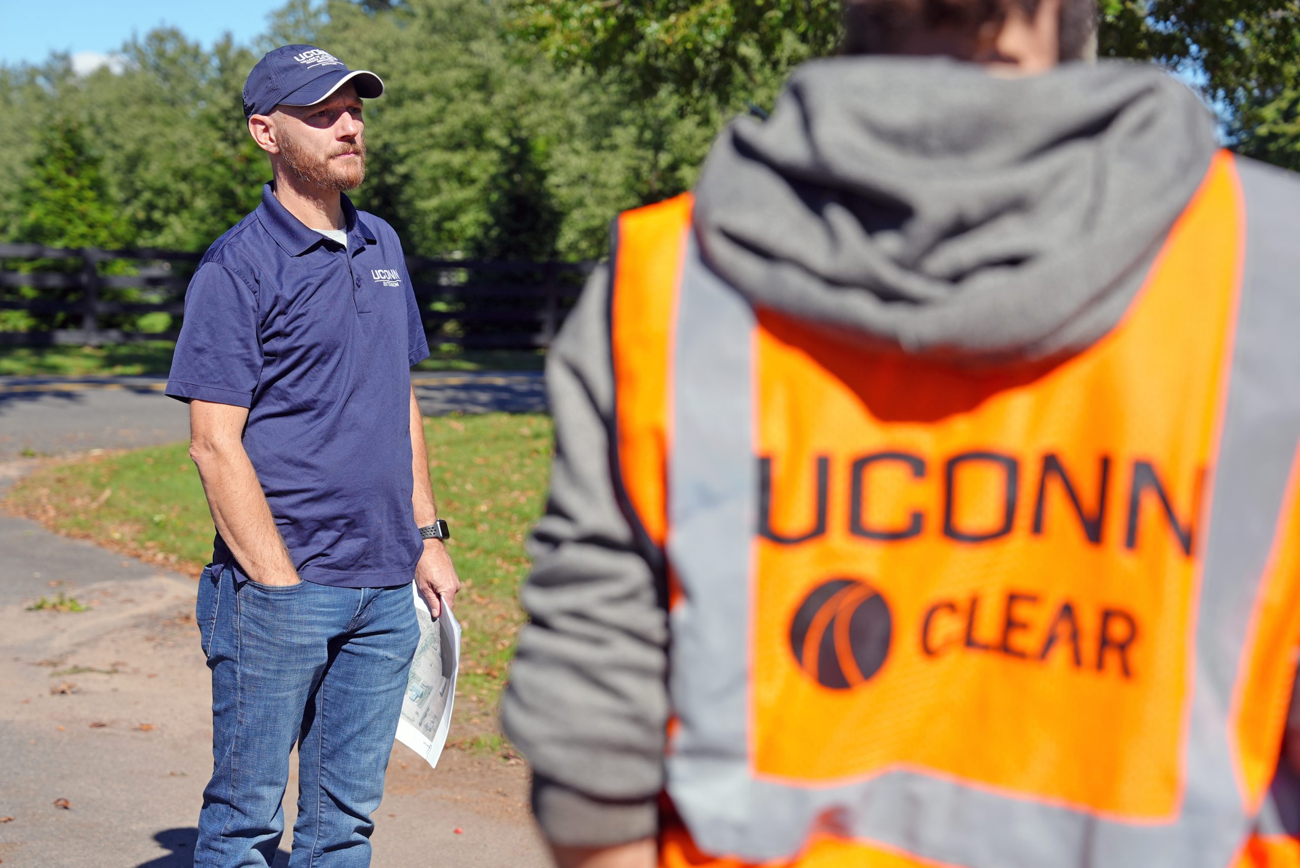 Two people standing outside, one in a blue shirt and a baseball cap, the other in a neon orange construction vest that says "UConn Clear"