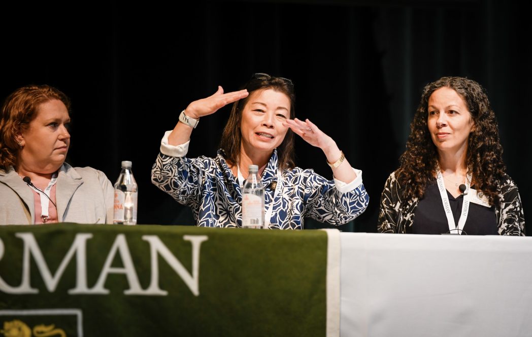 Three women sit at a table as the person in the middle gestures with her hands while talking.