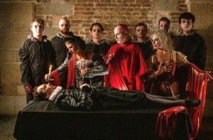 People dressed in black and red outfits stand over a reclined man presumably on his death