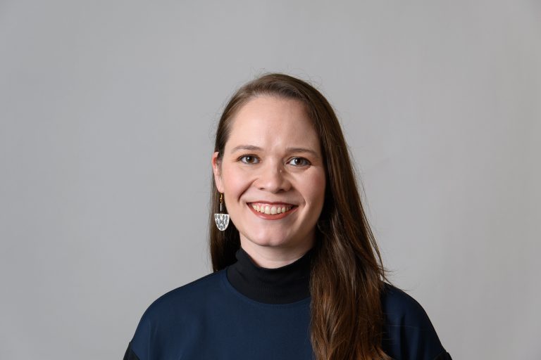 Headshot of a woman with long brown hair wearing a dark sweater and silver earrings against a grey background
