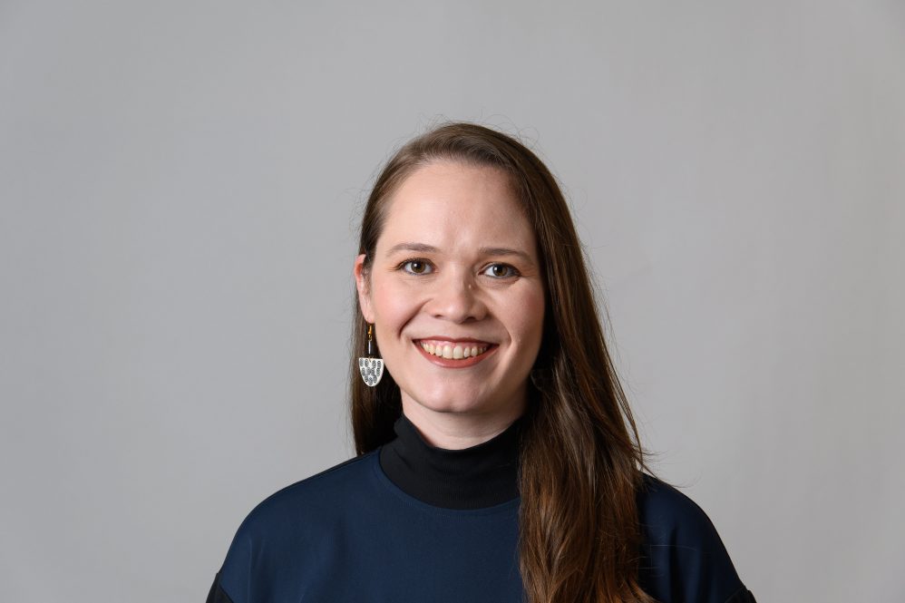 Headshot of a woman with long brown hair wearing a dark sweater and silver earrings against a grey background