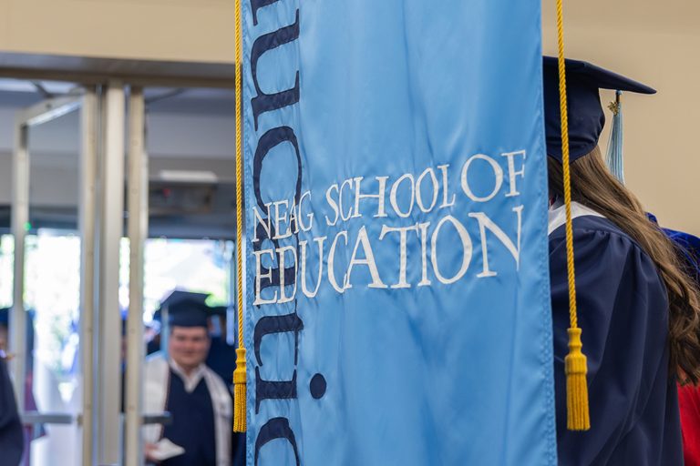 The UConn Neag School of Education banner is seen with graduates in the background.