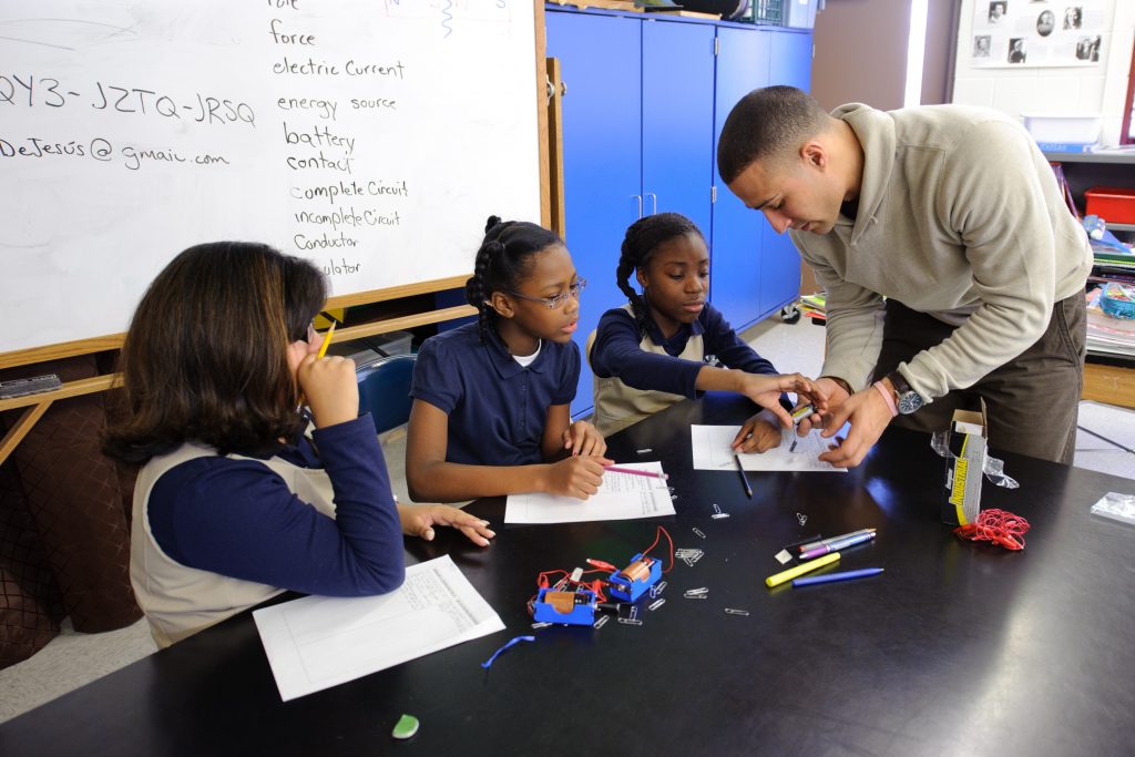 Three elementary school students working with their teacher to assemble circuits