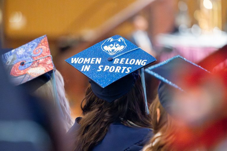 Graduates dressed in caps and robes sit in a row. One cap reads 
