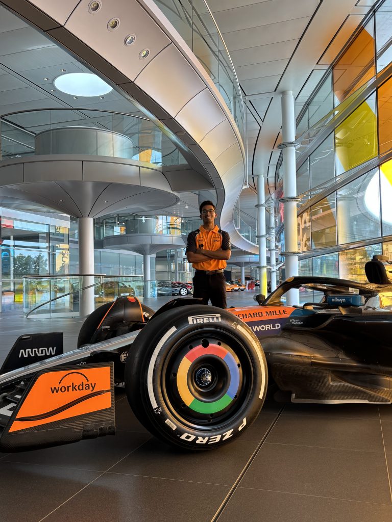 Engineer stands with arms crossed beside a McLaren Formula 1 car inside a modern showroom.