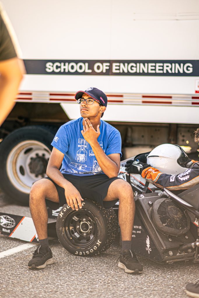 Engineering student sits beside a Formula SAE car, wearing a UConn shirt and cap.