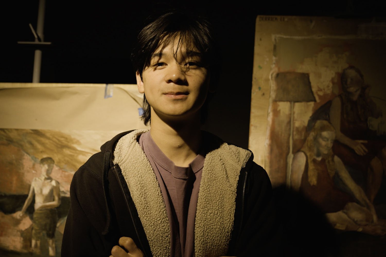 A man in a black sweatshirt and purple shirt stands in front of a pair of canvas oil paintings.