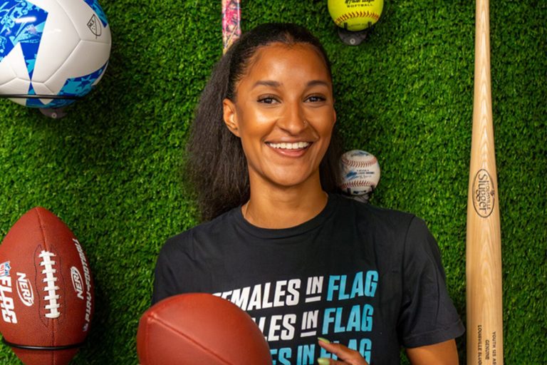 Jade Strawberry smiles and shifts a football in her hands. She wears a shirt that reads "Females in Flag". She is posed in front of a wall of green turf with baseball, baseball bat, soccer ball, football, and other sports gear.
