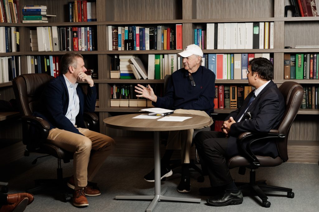 Three men sit at a table having a conversation, with book-lined shelves behind them.