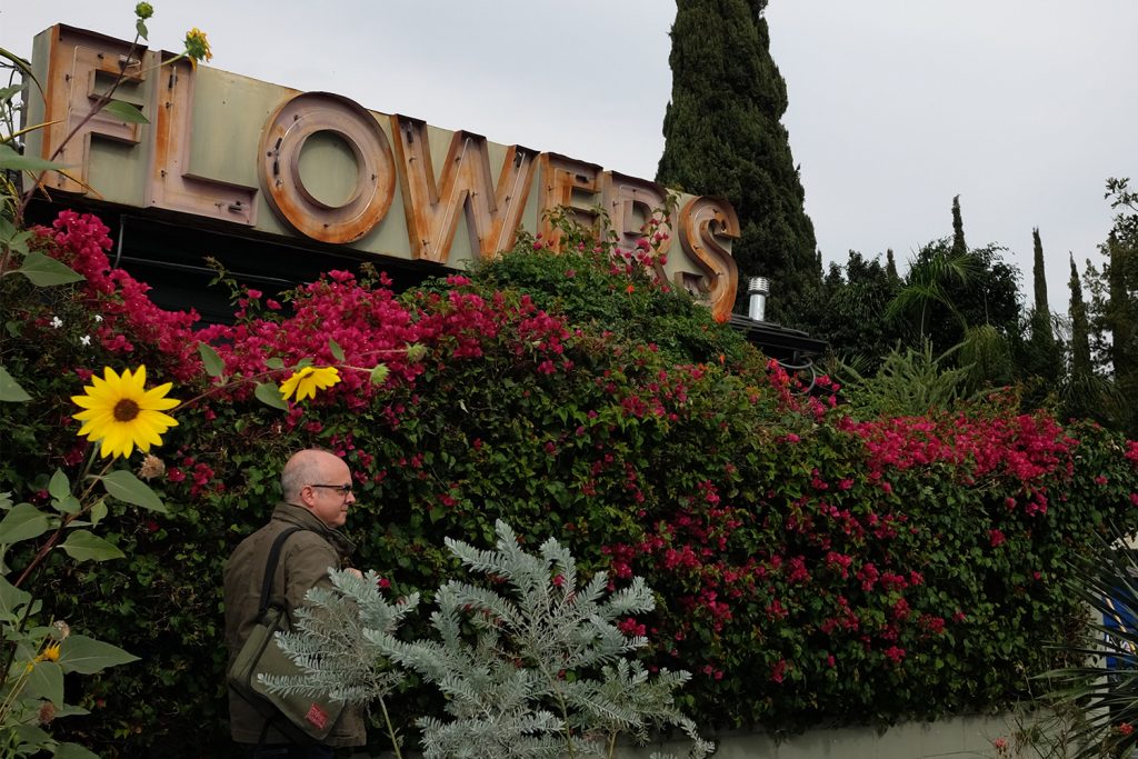 A man walking on a sidewalk with flowers and a sign that reads 'flowers' next to him